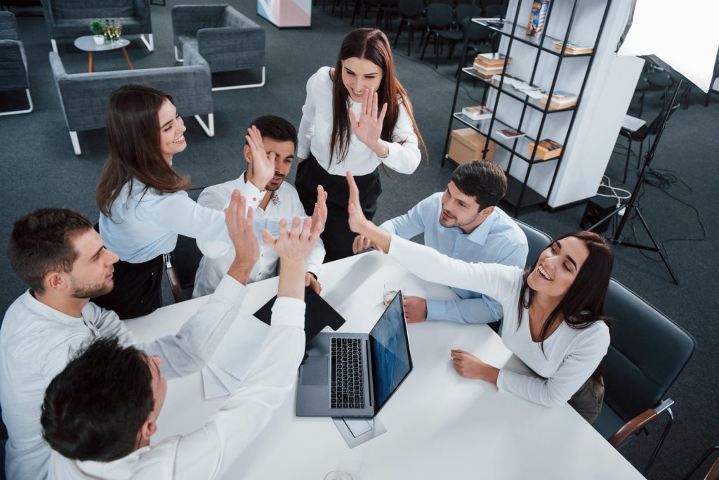 Seis personas con ropa de oficina chocan las manos alrededor de una mesa con un computador portátil. Imagen ilustrativa.